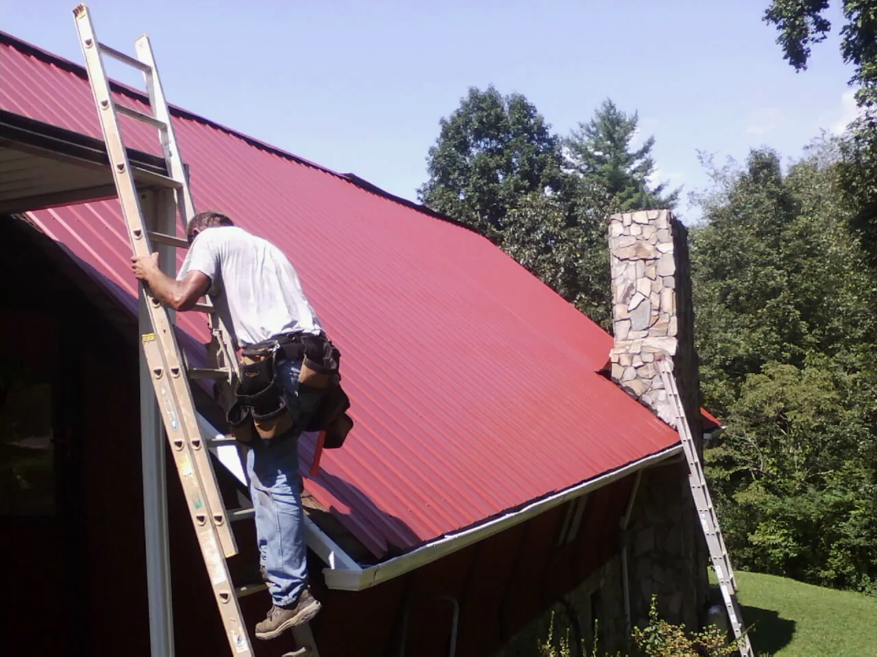Roofer on a ladder beside a red metal roof and stone chimney — Soffit & Fascia Repair by Roofing Brothers in Clarksdale